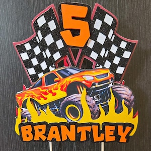 Puede incluir: Adorno para tarta de cumplea&ntilde;os con un monster truck en llamas, el n&uacute;mero 5 y banderas a cuadros. El nombre "BRANTLEY" se muestra en letras naranjas. El adorno est&aacute; sobre palitos de madera.