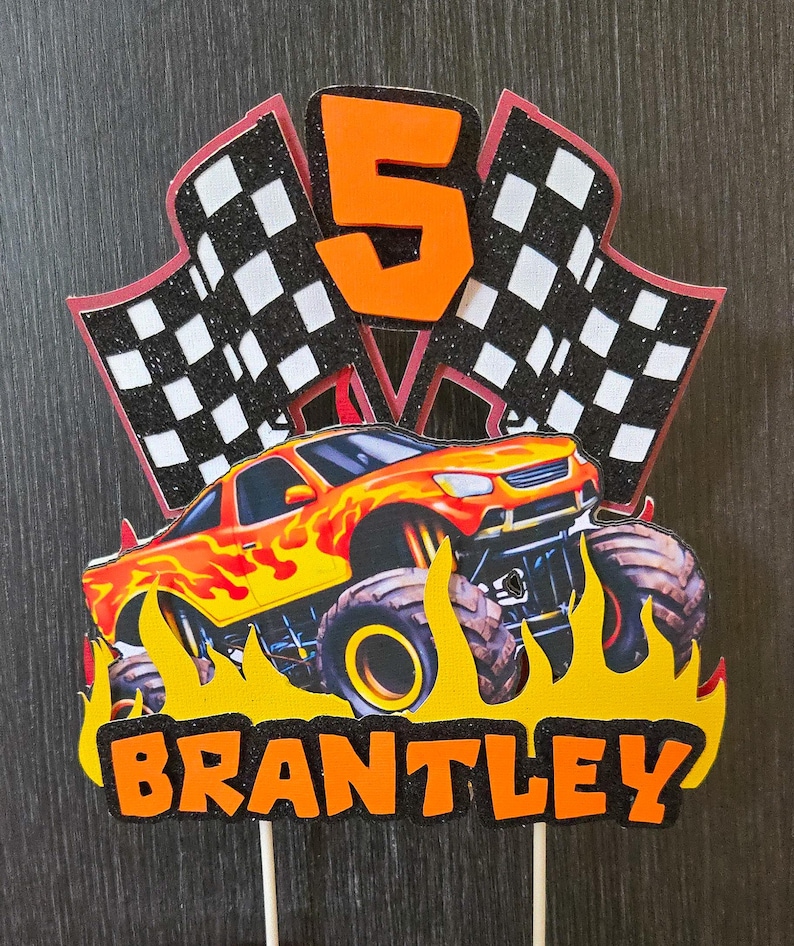 Puede incluir: Adorno para tarta de cumplea&ntilde;os con un monster truck en llamas, el n&uacute;mero 5 y banderas a cuadros. El nombre "BRANTLEY" se muestra en letras naranjas. El adorno est&aacute; sobre palitos de madera.