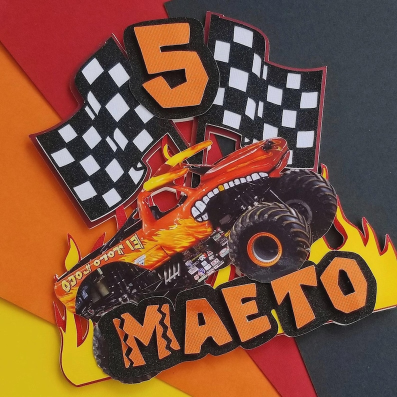 Puede incluir: Adorno de tarta de cumplea&ntilde;os colorido con un dise&ntilde;o de cami&oacute;n monstruo, el nombre "MAETO" y el n&uacute;mero "5". Incluye banderas a cuadros y llamas, con detalles en naranja, negro y blanco.