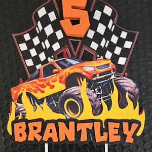 Puede incluir: Adorno de pastel colorido con un cami&oacute;n monstruo, llamas, banderas a cuadros y el n&uacute;mero 5. El nombre "BRANTLEY" se muestra en naranja. Ideal para una fiesta de cumplea&ntilde;os.