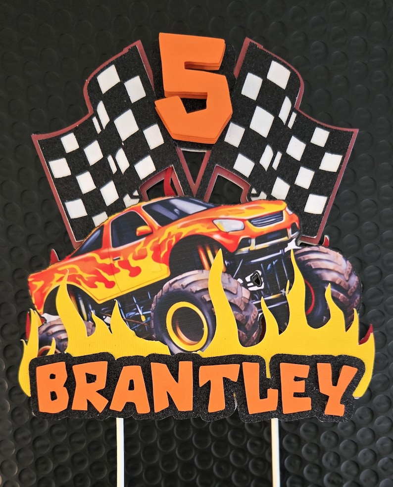 Puede incluir: Adorno de pastel colorido con un cami&oacute;n monstruo, llamas, banderas a cuadros y el n&uacute;mero 5. El nombre "BRANTLEY" se muestra en naranja. Ideal para una fiesta de cumplea&ntilde;os.