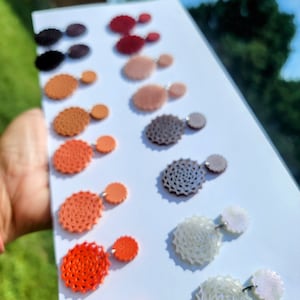 May include: A set of 10 pairs of earrings featuring a range of colours and designs. The earrings are crafted from polymer clay and have a circular shape. The colours vary from white to red, with shades of orange, pink, and brown in between. The earrings are displayed in rows on a white surface.