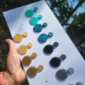 May include: A set of eight pairs of earrings featuring a layered, circular design. The earrings are available in a variety of colours, including teal, gold, black, and grey.