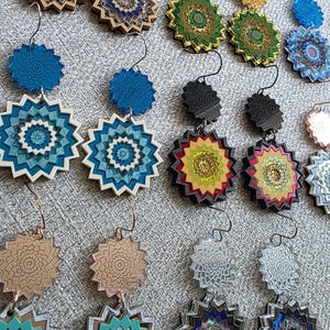 May include: A collection of layered, star-shaped dangle earrings featuring intricate geometric patterns. The earrings are crafted from various materials including metallic, translucent, and opaque acrylics in shades of blue, green, gold, and silver.