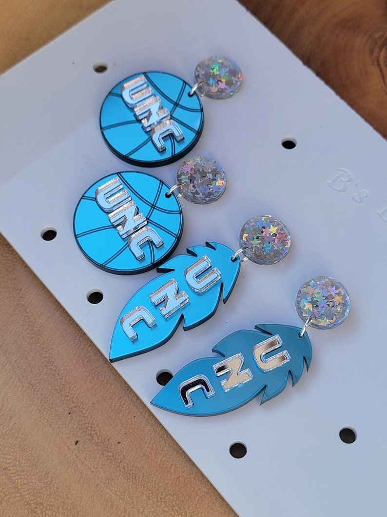 May include: A pair of earrings featuring a blue basketball with the word "UNC" in silver lettering and a blue feather with the word "UNC" in silver lettering. Each earring has a silver star-shaped charm.