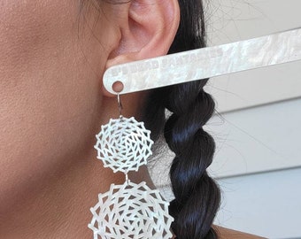 May include: White geometric dangle earrings with a layered, openwork design. The earrings are circular, with a smaller circle above a larger one. The earrings are suspended from silver-colored hooks. The image shows the earrings on a person.