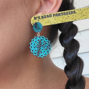 May include: A pair of turquoise and black earrings with a circular design. The earrings are made of wood and have a layered, geometric pattern. The earrings are shown being worn by a person with long black hair.