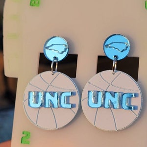 May include: A pair of blue and white basketball-themed earrings with a UNC logo and a North Carolina state outline.