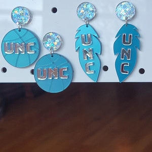 May include: A set of four UNC earrings. Two earrings feature a basketball design with the letters UNC in silver. The other two earrings feature a feather design with the letters UNC in silver.