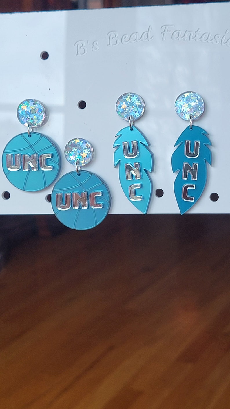 May include: A set of four UNC earrings. Two earrings feature a basketball design with the letters UNC in silver. The other two earrings feature a feather design with the letters UNC in silver.
