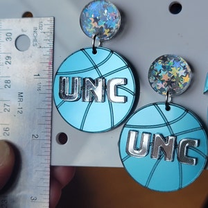 May include: A pair of blue and silver basketball-shaped earrings with a holographic star charm. The earrings feature the letters "UNC" in silver.