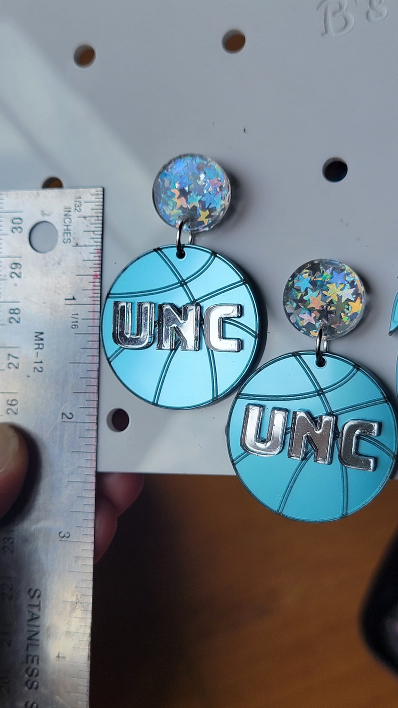 May include: A pair of blue and silver basketball-shaped earrings with a holographic star charm. The earrings feature the letters "UNC" in silver.