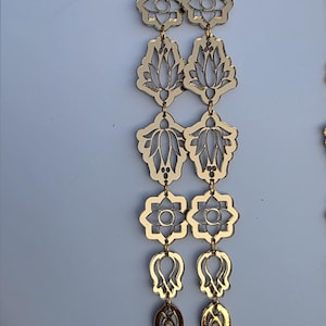 May include: Gold-colored, dangling earrings with a floral design. The earrings feature multiple connected shapes, including teardrops, flowers, and lotus-like petals. The earrings are displayed against a white background.