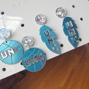 May include: A pair of earrings featuring a teal blue basketball with UNC written in silver lettering and a teal blue feather with UNC written in silver lettering. The earrings are accented with iridescent glitter.
