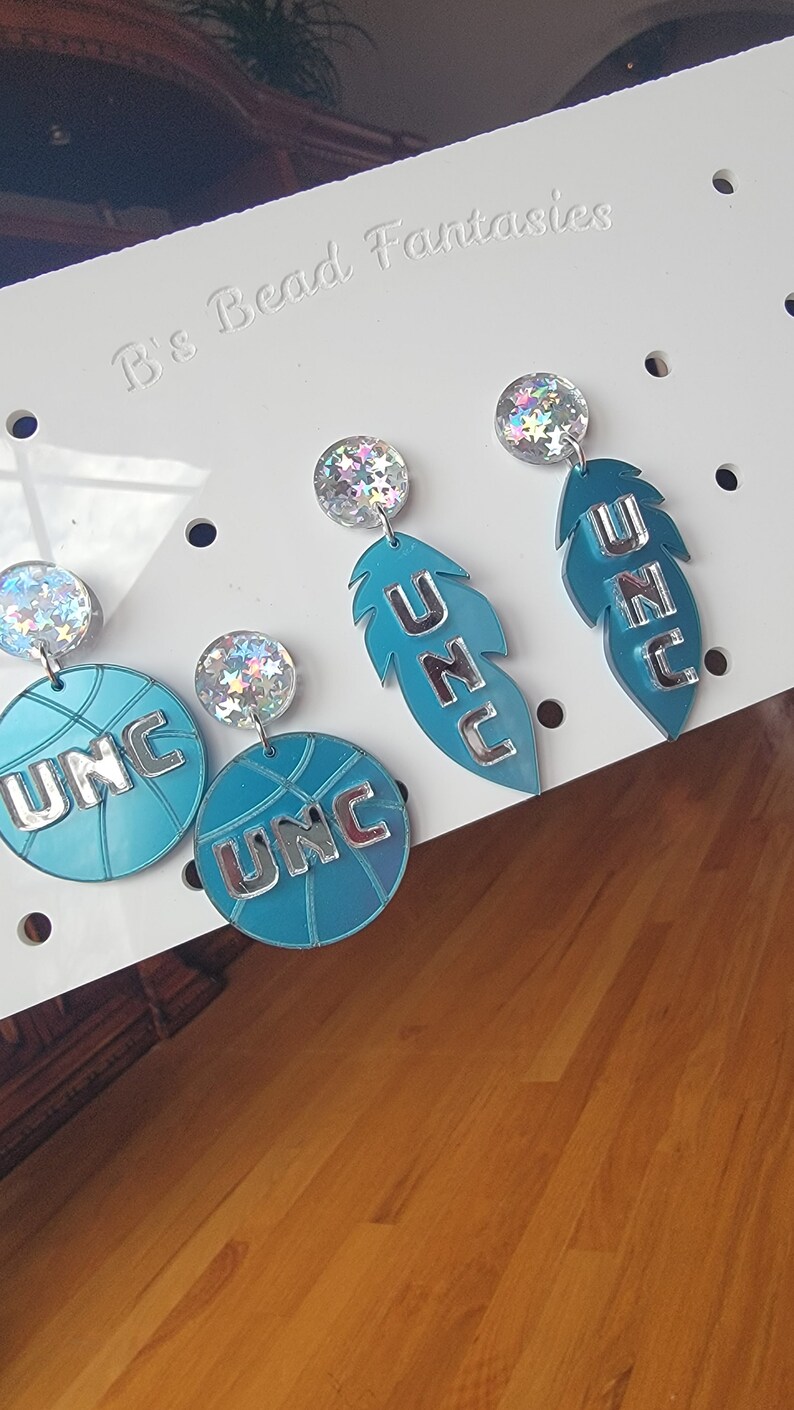 May include: A pair of earrings featuring a teal blue basketball with UNC written in silver lettering and a teal blue feather with UNC written in silver lettering. The earrings are accented with iridescent glitter.