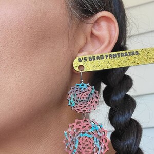 May include: A pair of dangle earrings with a geometric design. The earrings are made of metal and have a pink, blue, and silver color scheme. The earrings are hanging from a person's ear. The earrings are labeled "B's Bead Fantasies".