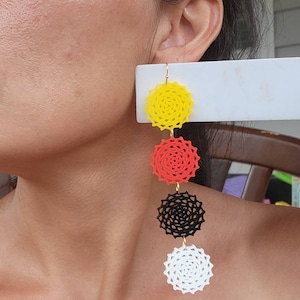 May include: Long, dangling earrings with four circular, geometric designs in yellow, red, black, and white. The earrings hang from a gold-coloured hook and a white rectangular backing. A striking, colourful accessory.