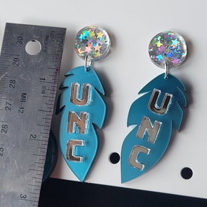 May include: A pair of blue feather-shaped earrings with silver lettering that reads "UNC". Each earring has a round iridescent charm with silver stars.