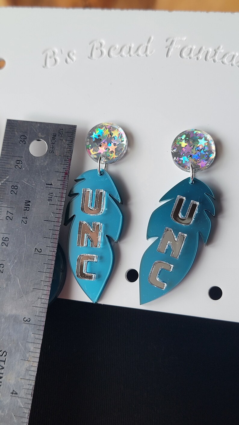 May include: A pair of blue feather-shaped earrings with silver lettering that reads "UNC". Each earring has a round iridescent charm with silver stars.