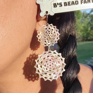 May include: A pair of white shell earrings with a geometric design. The earrings are hanging from a person's ear and are attached to a silver hook. The earrings are labelled "B's Bead Fantasy".
