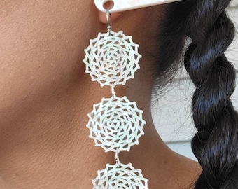 May include: A pair of long, silver earrings with four white, circular, openwork designs. The earrings are hanging from a person's ear. The text "BIG BEAD FANTASIES" is visible on a white, rectangular object.