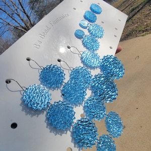 May include: A set of blue beaded earrings with a circular design. The earrings are displayed on a white board with the text "B's Bead Fantasies".