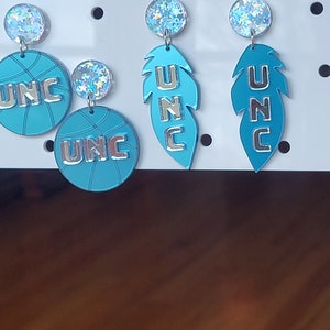 May include: A set of three pairs of earrings featuring a basketball, a feather, and a feather with the letters UNC in silver on a blue background.