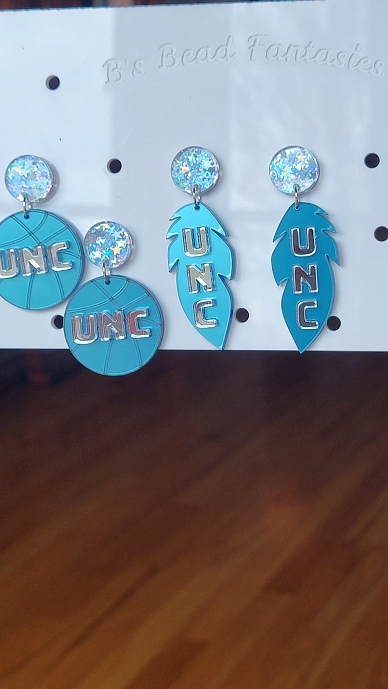 May include: A set of three pairs of earrings featuring a basketball, a feather, and a feather with the letters UNC in silver on a blue background.
