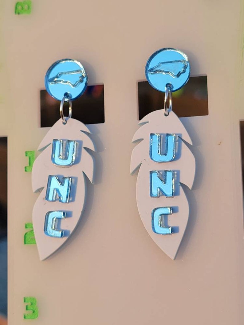 May include: A pair of white feather earrings with blue UNC lettering and a blue North Carolina state outline charm.