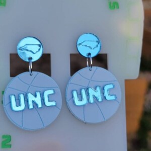 May include: A pair of blue and white basketball-themed earrings with a North Carolina state outline charm. The earrings feature the letters "UNC" in blue.