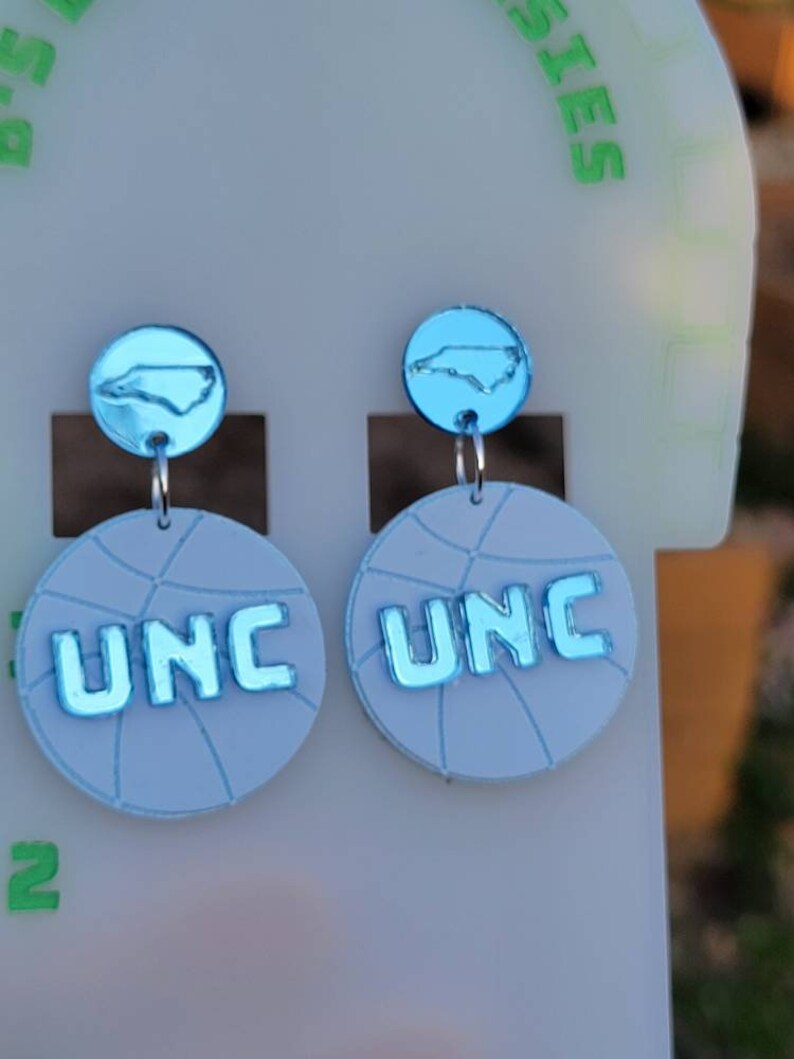 May include: A pair of blue and white basketball-themed earrings with a North Carolina state outline charm. The earrings feature the letters "UNC" in blue.