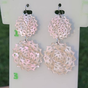 May include: A pair of white, double-layered earrings with a delicate, intricate design. The earrings are made of a white material with a subtle iridescent sheen. The earrings are suspended from silver-toned hooks.