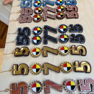 May include: A collection of decorative number and symbol ornaments in various colors and materials. The ornaments include the numbers 5 and 7, along with a sun-like symbol. Materials include glitter, wood grain, and metallic finishes.