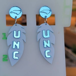 May include: A pair of blue and white dangle earrings featuring a North Carolina state outline charm and a feather charm with the letters UNC.
