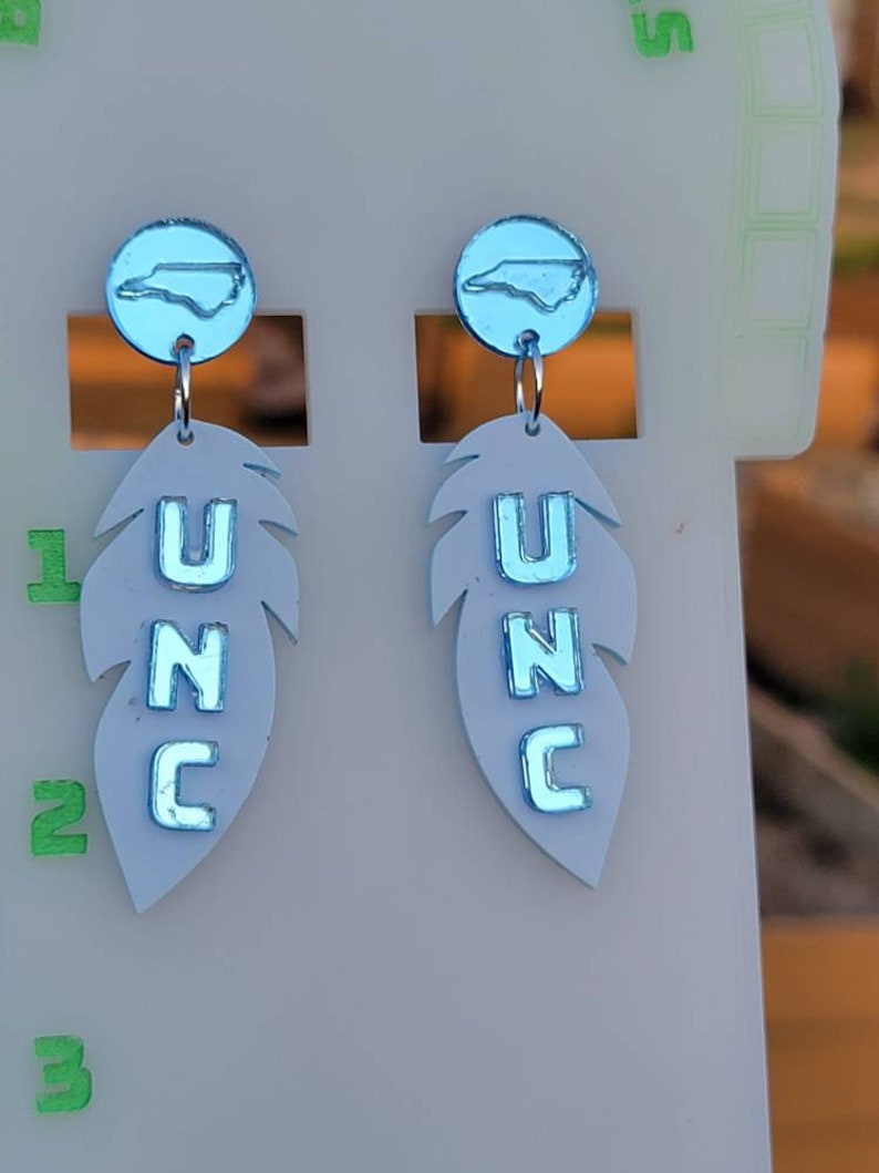 May include: A pair of blue and white dangle earrings featuring a North Carolina state outline charm and a feather charm with the letters UNC.