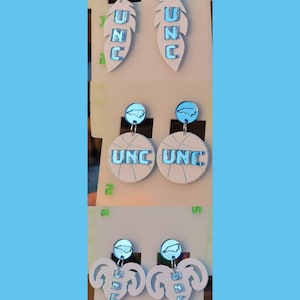 May include: Three pairs of earrings featuring a UNC logo. The first pair features a white feather with a blue UNC logo. The second pair features a white basketball with a blue UNC logo. The third pair features a white heart with a blue UNC logo.