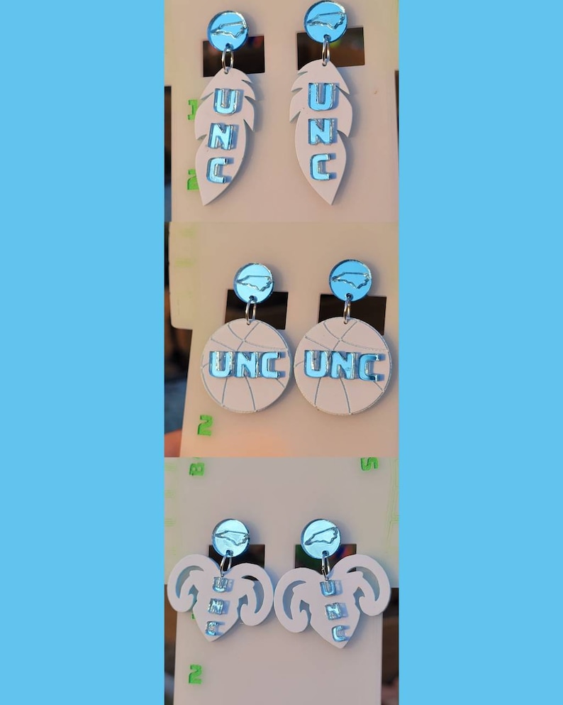 May include: Three pairs of earrings featuring a UNC logo. The first pair features a white feather with a blue UNC logo. The second pair features a white basketball with a blue UNC logo. The third pair features a white heart with a blue UNC logo.