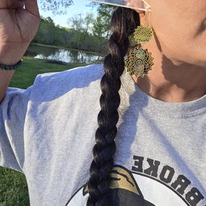 Puede incluir: Pendientes colgantes geom&eacute;tricos de color dorado con un dise&ntilde;o en capas. Los pendientes se llevan con una sudadera gris. Se sostiene una regla transparente con el texto "Read Fantasies" cerca de la oreja. Se ve una larga trenza oscura.