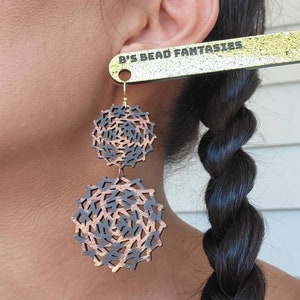 May include: A pair of dangle earrings with a black and brown geometric pattern. The earrings are made of wood and have a gold-tone finish. The earrings are hanging from a person's ear. The text "B's Bead Fantasies" is visible on a gold glitter background.