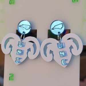 May include: A pair of white earrings shaped like a ram's head with a blue North Carolina state outline charm hanging from each earring. The charms are labelled "UNC" in blue letters.