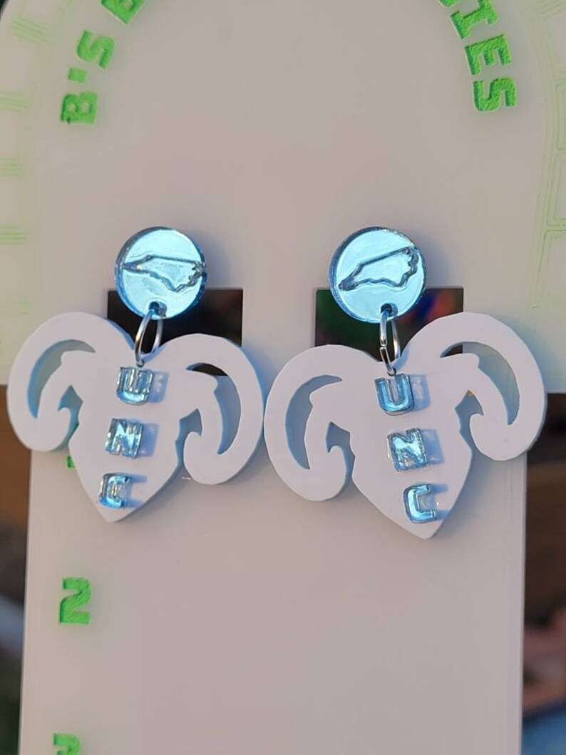 May include: A pair of white earrings shaped like a ram's head with a blue North Carolina state outline charm hanging from each earring. The charms are labelled "UNC" in blue letters.