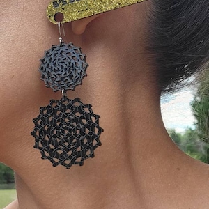 May include: A pair of black geometric earrings with a circular design. The earrings are hanging from a person's ear. The text "B's Bead Fantasies" is visible on a gold glitter background.