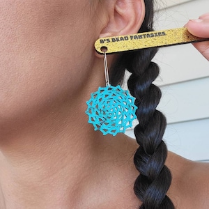 May include: A pair of turquoise blue earrings with a circular, geometric design. The earrings are hanging from a silver post and are being held up by a wooden ruler with the text "B's Bead Fantasies" printed on it.
