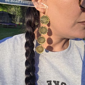 Puede incluir: Pendientes dorados en capas con dise&ntilde;o floral. Los pendientes cuelgan de la oreja de una persona que lleva una camiseta gris. Los pendientes tienen forma circular con intrincados patrones calados. El texto "Bs Bead Fantas" es visible.
