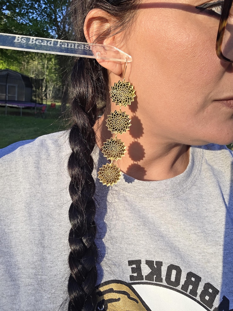 Puede incluir: Pendientes dorados en capas con dise&ntilde;o floral. Los pendientes cuelgan de la oreja de una persona que lleva una camiseta gris. Los pendientes tienen forma circular con intrincados patrones calados. El texto "Bs Bead Fantas" es visible.