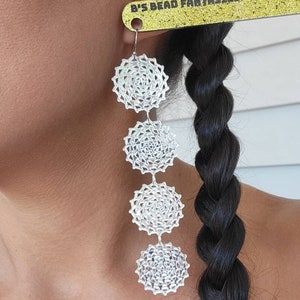 May include: Silver-coloured, dangling earrings with a layered, circular design. The earrings feature four connected, starburst-shaped elements. The text "B'S BEAD FANTASIES" is visible on a gold-coloured tag attached to the earring.