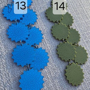 Puede incluir: Dos pares de pendientes colgantes. Un par es azul vibrante, el otro verde oliva. Cada pendiente presenta una serie de colgantes conectados en forma de estrella con intrincados patrones circulares. Los pendientes se muestran sobre una superficie texturizada de color gris claro. Los n&uacute;meros 13 y 14 est&aacute;n en la imagen.
