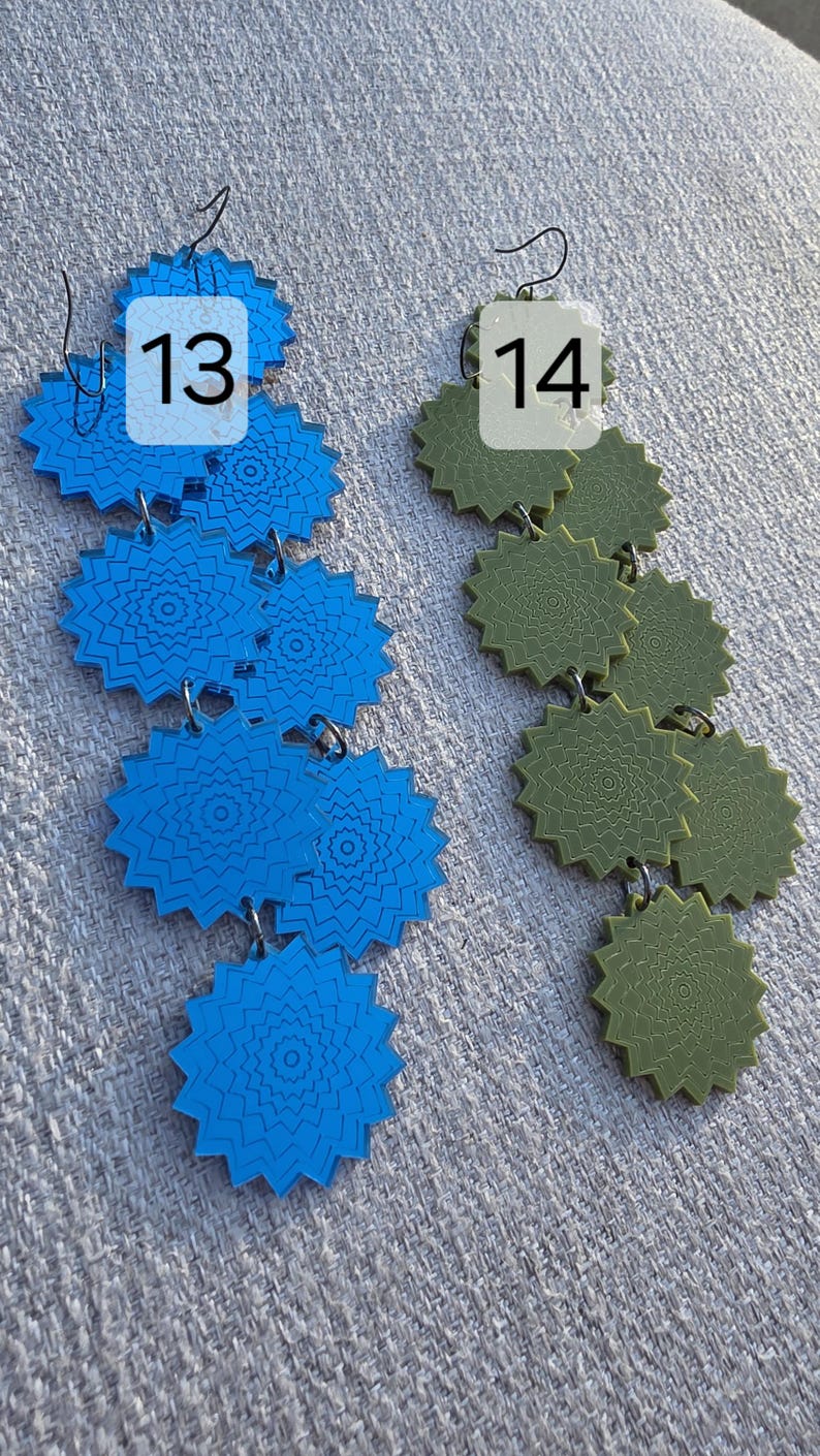 Puede incluir: Dos pares de pendientes colgantes. Un par es azul vibrante, el otro verde oliva. Cada pendiente presenta una serie de colgantes conectados en forma de estrella con intrincados patrones circulares. Los pendientes se muestran sobre una superficie texturizada de color gris claro. Los n&uacute;meros 13 y 14 est&aacute;n en la imagen.
