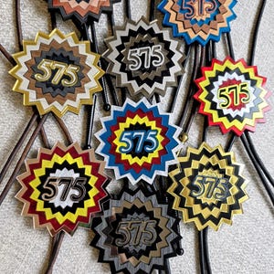 May include: A collection of bolo ties with starburst-shaped pendants. Each pendant features the number "575" in a contrasting color. The pendants are in various colors, including brown, blue, gold, and red, with black cords.