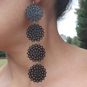 May include: Black dangle earrings with four circular, lacy, black metal charms hanging from a silver hook. The earrings are shown on a person's ear.